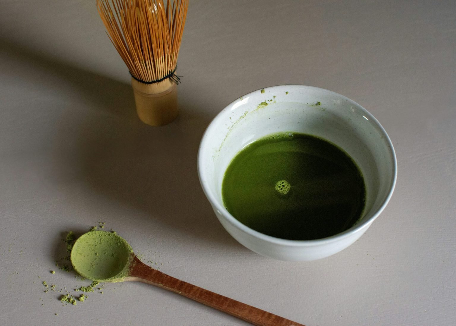 A white bowl of whisked matcha beside a bamboo chasen and a wooden scoop of powder