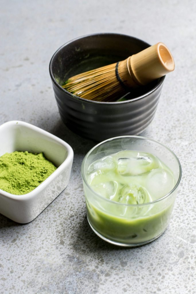 Matcha powder, a black chawan and an iced matcha on a grey surface