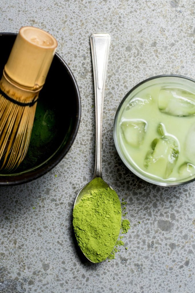 A spoon of matcha powder beside a bowl and whisk, shot from above